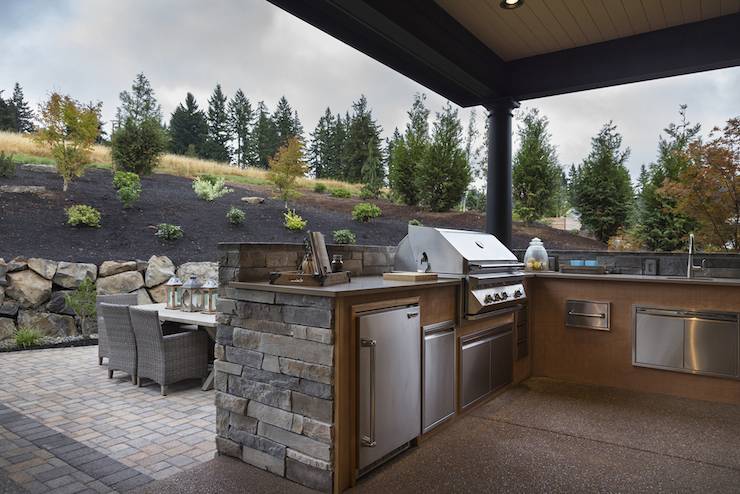 Stunning covered patio features an L shaped outdoor kitchen clad in stacked stone fitted with a stainless steel mini fridge alongside an integrated barbecue and outdoor sink framed by brown countertops across from a brick paver patio which highlights an outdoor trestle dining table lined with woven outdoor dining chairs.