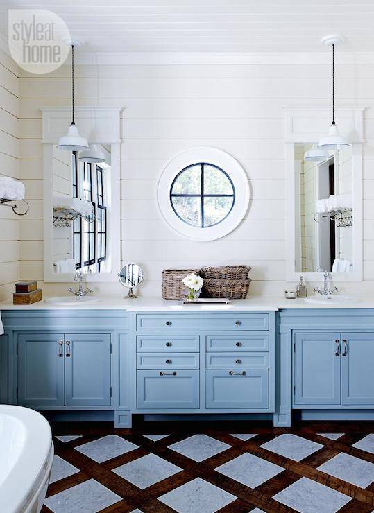 Beautiful coastal bathroom with built-in sky blue dual sink vanity accented with nickel pulls alongside white counters which frame oval porcelain sinks situated below white vanity mirrors on tongue and groove clad walls lit by white enamel pendants on either side of a round window. The blue double vanity stands atop an inset wood trellis patterned floors framing marble tile laid on the diagonal.