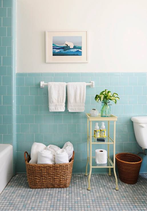 Pretty bathroom with aqua blue tiled half walls and bath surround. The blue from the tile is picked up in the coastal print hanging on the ivory painted wall above. Mosaic tiles in varying shades of blue cover the floor and add to the watery color palette. A large seagrass basket holds rolled towels whilst a pale yellow metal and glass etagere holds flowers and other bath essentials.