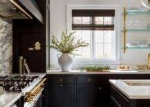 Stylish black kitchen boasts black cabinets donning brass knobs and holding a sink with a brass gooseneck faucet beneath glass and brass shelves.