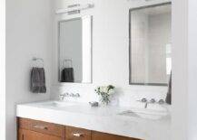 Bathroom features a brown wooden double vanity topped with white marble under nickel mirrors, illuminated by long white glass sconces and light gray floor tiles.