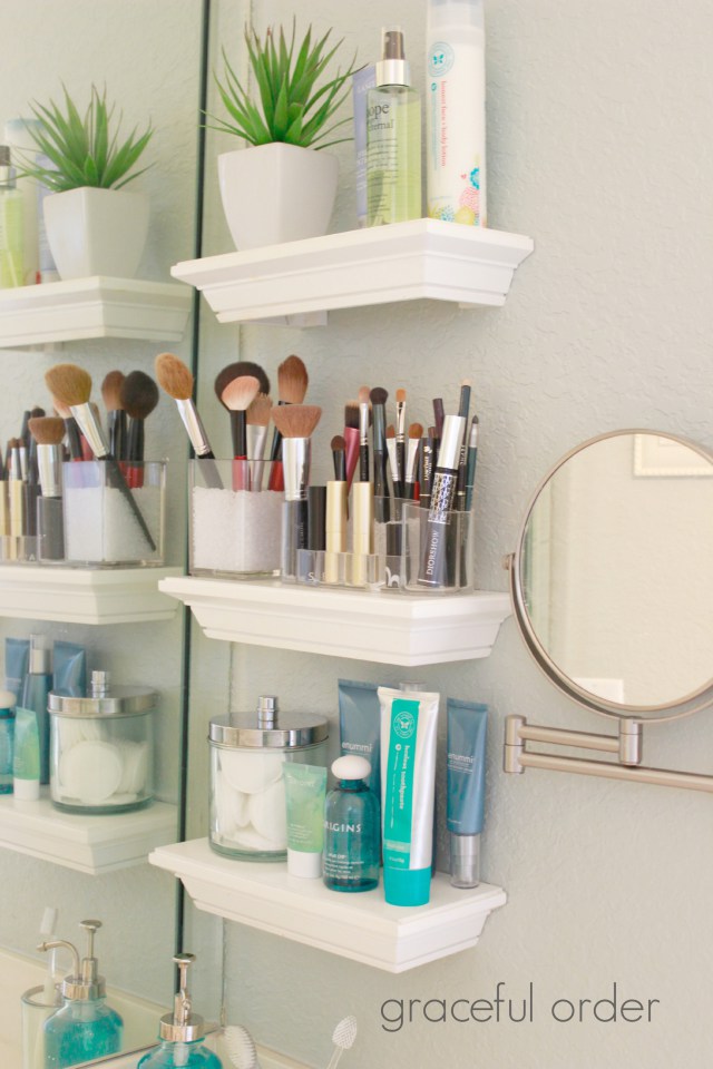 Three small white floating shelves in a bathroom that carry makeup brushes and skin care products.