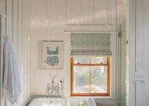 Charming white and blue cottage bathroom features a sky blue clawfoot bathtub placed on blue mosaic floor tiles in front of a polished nickel floor mount tub filler and beneath a framed blue swimsuit art piece hung from a board and batten wall. The room is illuminated by a mint green vintage lantern.