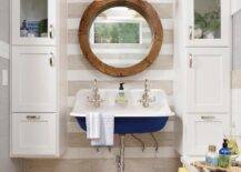 A window framed by white and gray striped wall tiles is located over a round wooden mirror hung between glass front cabinets stacked over white linen cabinets. A blue Kohler Brockway Sink floats over river rock floor tiles. A white built-in bench boasts cubbies stuffed with towels.