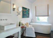 Tranquil bathroom with acrylic freestanding bath tub from Giagni with polished nickel floor-mount tub filler. Bathroom window covered in white roman shade with tan ribbon trim and glossy espresso hardwood bathroom floors. Polished nickel sconces flanking Restoration Hardware Hutton Mirror over white bathroom vanity with white vessel sink and wall-mount faucet kit. Bathroom with Global Views Directoire Table and white slipcovered armchair.