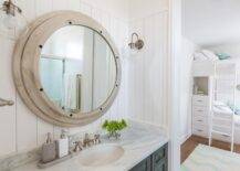 Kids' cottage bathroom features vertical shiplap walls lined with a large round nautical mirror illuminated by clear glass barn sconce over a gray distressed washstand topped with white marble fitted with an oval sink and satin nickel faucet.