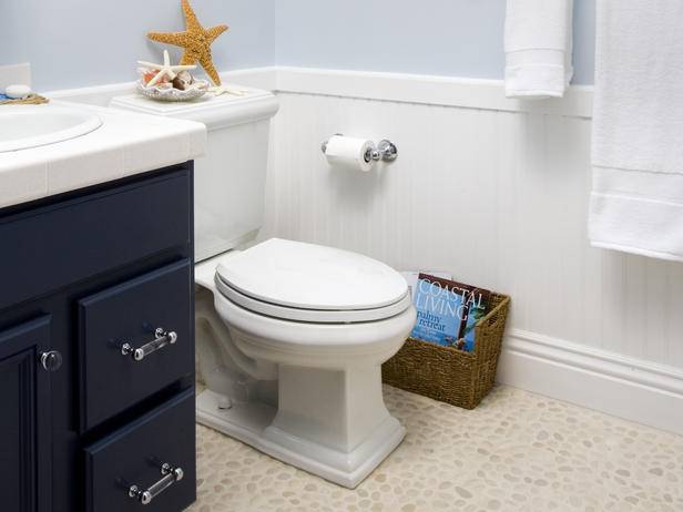 Coastal style blue bathroom with pale blue walls paired with white beadboard surround. Navy blue bathroom vanity with nickel and acrylic hardware. Beautiful pebble bathroom floor, wicker magazine rack and beachy accents.