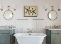 Cottage kids' bathroom features upper walls painted a creamy white and lower walls clad in vertical shiplap lined with a freestanding tub paired with a vintage style tub filler under side by side towel rails under starfish art flanked by gray wash washstands. Shared kids' bathroom boasts separate gray distressed bathroom vanities topped with marble countertops under Arteriors Lander Iron Mirrors illuminated by polished nickel single sconces.