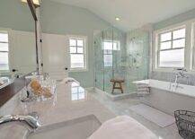 Spa like bathroom features a carrera top dual vanity fitted with his and hers sinks under a full length espresso framed mirror alongside a gray marble tiled floor set in a diamond pattern. A freestanding tub, placed below two windows, stands next to a corner shower clad in grey marble tiles accented with white linear glass tiles.