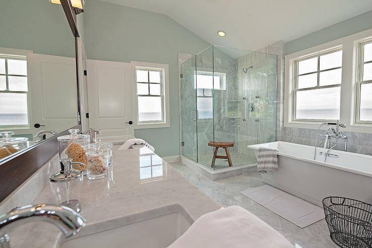 Spa like bathroom features a carrera top dual vanity fitted with his and hers sinks under a full length espresso framed mirror alongside a gray marble tiled floor set in a diamond pattern. A freestanding tub, placed below two windows, stands next to a corner shower clad in grey marble tiles accented with white linear glass tiles.