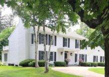 A circular driveway leads to a white brick colonial home is contrasted with black shutters and accented with a portico finished with Greek columns lit by carriage lanterns.