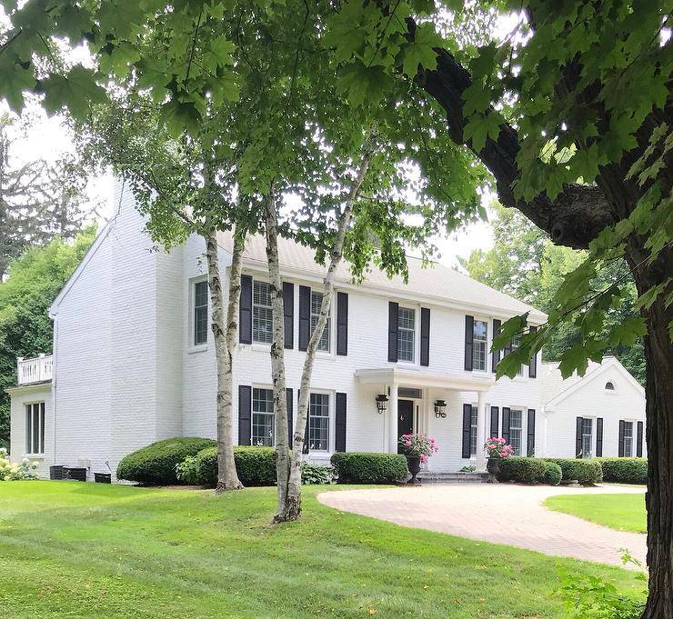 A circular driveway leads to a white brick colonial home is contrasted with black shutters and accented with a portico finished with Greek columns lit by carriage lanterns.