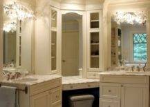 Beautiful master bathroom design with gray vanity ottoman tucked under corner vanity accented with inset mirror and glass-front cabinets flanked by his and her vanities featuring ivory cabinets paired with white marble countertops and sconces mounted on inset mirrors.