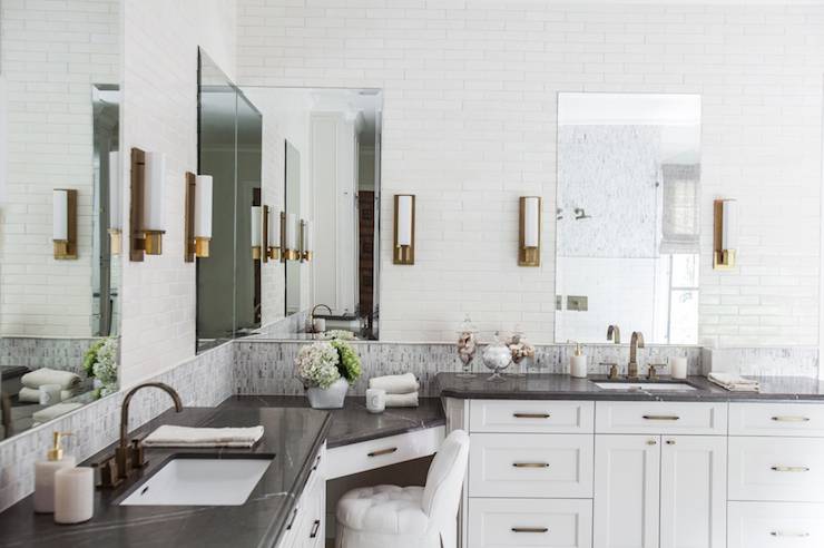 Beautiful black and white bathroom features his and her vanities paired with soapstone countertops framing sinks accented with brass gooseneck faucets and mosaic tile backsplash flanking drop down corner vanity paired with white tufted vanity stool under wraparound mirror. Frameless mirrors illuminated by brass sconces mounted on ceiling height off-white linear tiled backsplash over each sink.