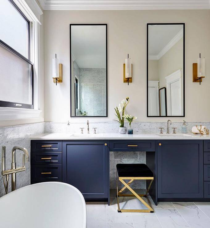 Bathroom features tall black vanity mirrors flanked by brass and white sconces over a gray marble vanity backsplash and a blue double washstand with brass pulls, a gray marble countertop with a sink that boasts polished nickel faucets and a gold and black vanity stool.