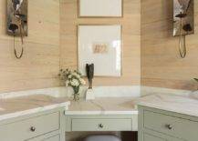 Two gold framed art pieces hang from a tan plank wall over a green corner makeup vanity topped with a honed white marble countertop paired with a gray counter stool placed on gray floor tiles. The vanity is flanked by green washstands accented with satin nickel knobs and lit by mirrored sconces.