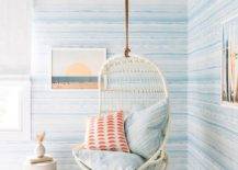 Charming white and blue bedroom features a white rattan hanging chair topped with a blue chevron and orange pillow and hung in a corner in front of walls coered in blue striped wallpaper.