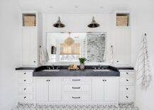 White and gray lattice floor tiles lead to a white plank double washstand adorned with oil rubbed bronze hardware and a thick honed black marble countertop. Sink with polished nickel faucet are located under a frameless vanity mirror lit by two vintage polished nickel lanterns mounted to a vertical plank backsplash. The lanterns are flanked by cubbies holding woven bins placed over linen cabinets.