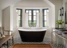 Cottage style bathroom features a bay window with black clawfoot tub on black and white mosaic floor tiles, a black top sink vanity and a wooden bench.