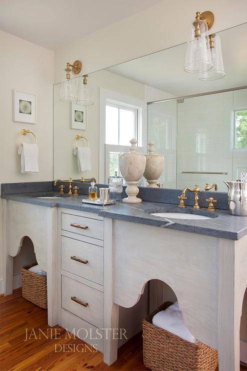 Glass and brass sconces are mounted over a full length frameless vanity mirror hung over antique brass hook and spout faucets mounted to a black honed marble countertop accenting a gray brushed oak dual washstand.