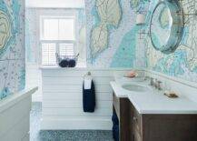 Brown and blue boy's cottage bathroom features a brown washstand fitted with a white quartz countertop finished with an oval sink with a polished nickel faucet beneath a porthole mirror mounted between Thomas O'Brien Bryant Sconces on a wall covered in world map wallpaper accented with a shiplap trim as blue hexagon floor tiles cover the floor.