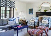 Mixed patterns, color schemes, and design furnishings come together beautifully to create a contemporary living space. A blue denim sectional with nailhead trim is decked with a few blue chinoiserie pillows next to a blue Charleston Coffee Table on a light blue area rug. White and blue geometric print chairs mismatch next to colorful striped x stools but manage to contrasts one another in a bold yet fitting combo. Black wooden fireplace mantle with a brass arabesque screen gives a platform for a lovely large gold mirror flanked by two floral arrangements in a white square vase. A brass and cream Aspect Sconce is mounted next to a black framed window dressed in blue and white pattern shade over a round wood end table featuring a white double gourd lamp.