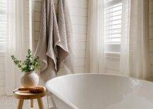 Modern brass towel hooks are fixed to a shiplap wall in a cottage bathroom featuring windows covered in white shutters and sheer white curtains.