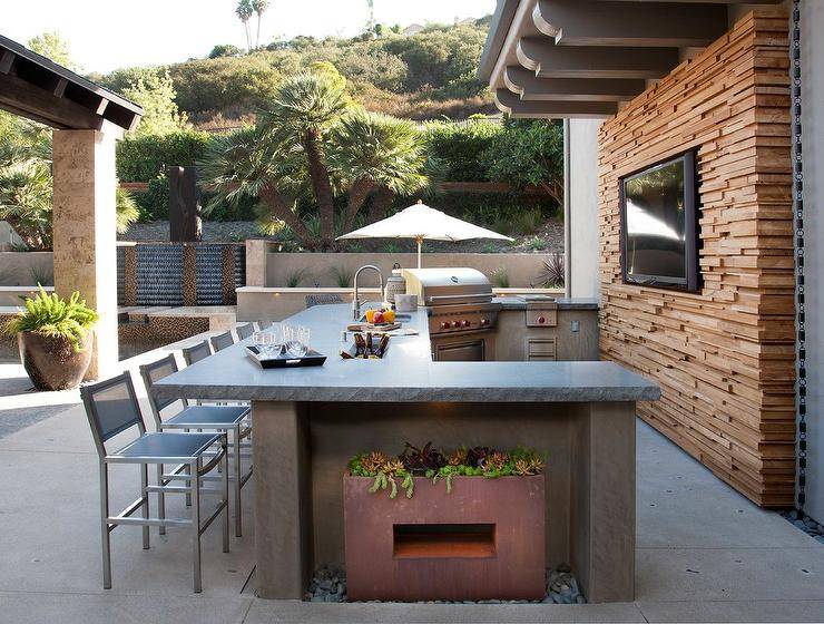 U shaped outdoor kitchen boasts concrete countertops fitted with a sink with polished nickel gooseneck faucet and a stainless steel barbecue. Stainless steel barstools sit facing a flank accent wall finished with an inset flat panel television.