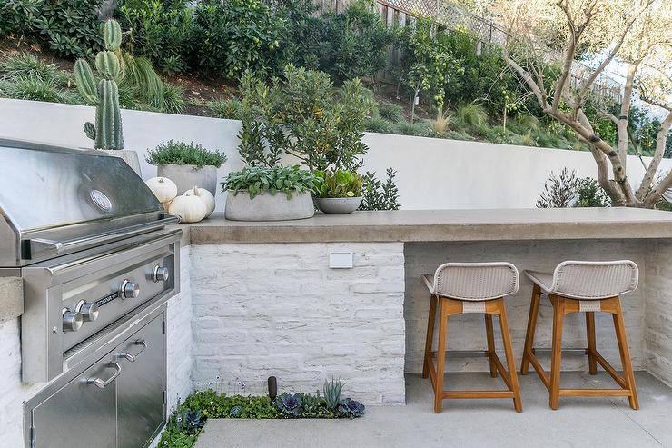 Low back gray rattan barstools sit beneath a concrete countertop accenting a l-shaped outdoor kitchen finished with a stainless steel barbecue.