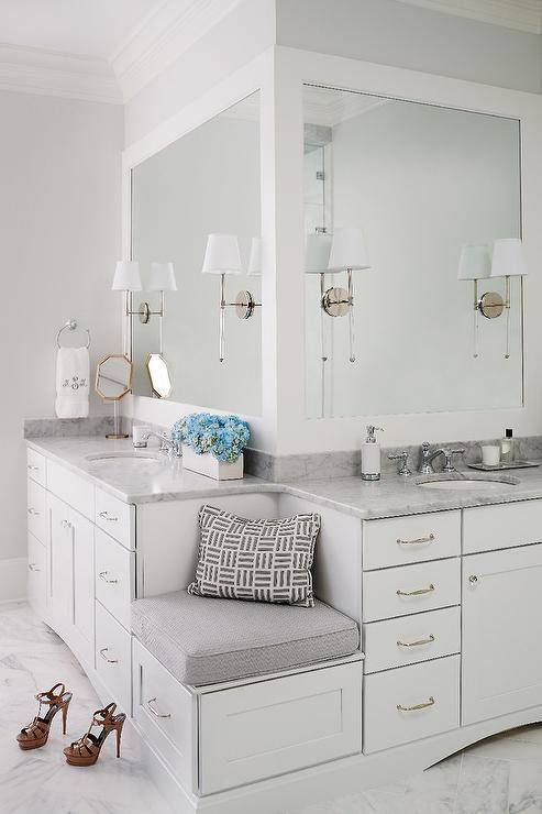 A built-in corner bench fitted with a single drawer adorned with a polished nickel pull topped with a gray cushion is flanked by his and her washstands topped with carrera marble placed under Camille Long Sconces mounted on thick, white framed vanity mirrors.