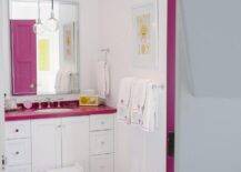 A hot pink door opens to a contemporary pink and white girl's bathroom featuring a white washstand finished with nickel hardware and a hot pink countertop holding an oval sink with a polished nickel faucet beneath a polished vanity mirror mounted on a white wall lit by light bulb pendant. A white step stool sits on a hot pink striped bath mat on gray wood-like floor tiles in front the washstand while a white and yellow framed print hangs above a polished nickel towel bar.