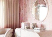 Charming contemporary bedroom features walls covered in pink wallpaper and a "Dream of Me" mirror hung above an oblong credenza, as a light pink velvet accent chair sits in a corner on a cream rug.