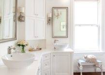 White bathroom features curved cabinets topped with his and her bowl sinks paired with off-set faucets and beveled mirrors flanking corner center console cabinet accented with antiqued sconces illuminated by crystal chandelier.