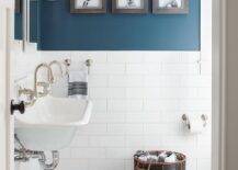 Well appointed blue and white boy bathroom boasts walls clad Benjamin Moore Newburg Green paint accented with large white subway backsplash tiles complementing white hexagon floor tiles. Three framed animal prints are mounted above a black wicker laundry basket on a wall adjacent to a white beveled vanity mirror mounted between bell jar sconce above a Kohler Brockway Sink fitted with gooseneck faucets and kid-accessible via a Serena & Lily Teak Step Stool.