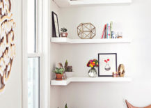 Three white corner-mounted floating shelves hold a small plant, pictures, and a uniquely shaped decor piece.